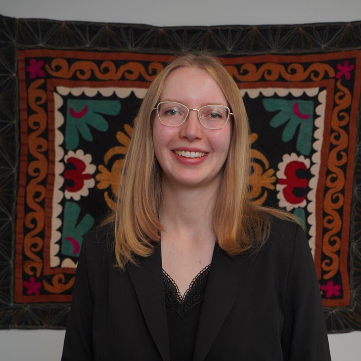 A woman with long, straight hair and glasses stands in front of a vibrant tapestry featuring symmetrical floral and geometric patterns in red, green, white, and orange on a black background. They are wearing a black blazer over a lace-trimmed top and smiling at the camera.