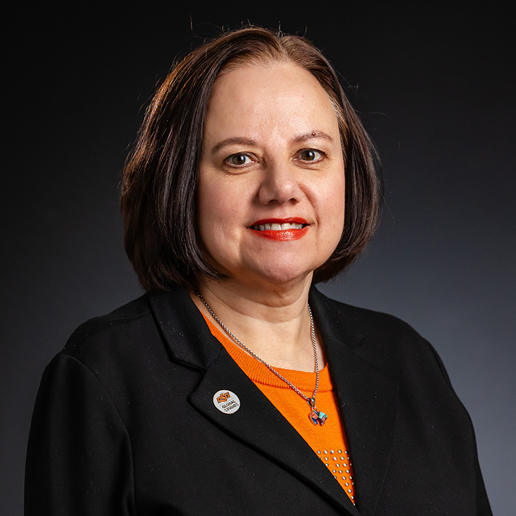 A person with short dark hair wearing a black blazer over an orange top, accessorized with a butterfly pendant necklace. A round pin on the blazer reads 'OSU College of Education.' The background is dark and plain.