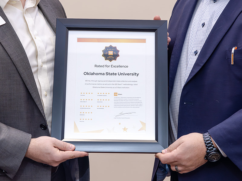 Two individuals hold a framed certificate that reads 'Rated for Excellence, Oklahoma State University.' The certificate features a badge at the top and indicates a 5 Stars Institution rating by QS Stars. It includes multiple star ratings, detailed text, and a signature at the bottom.