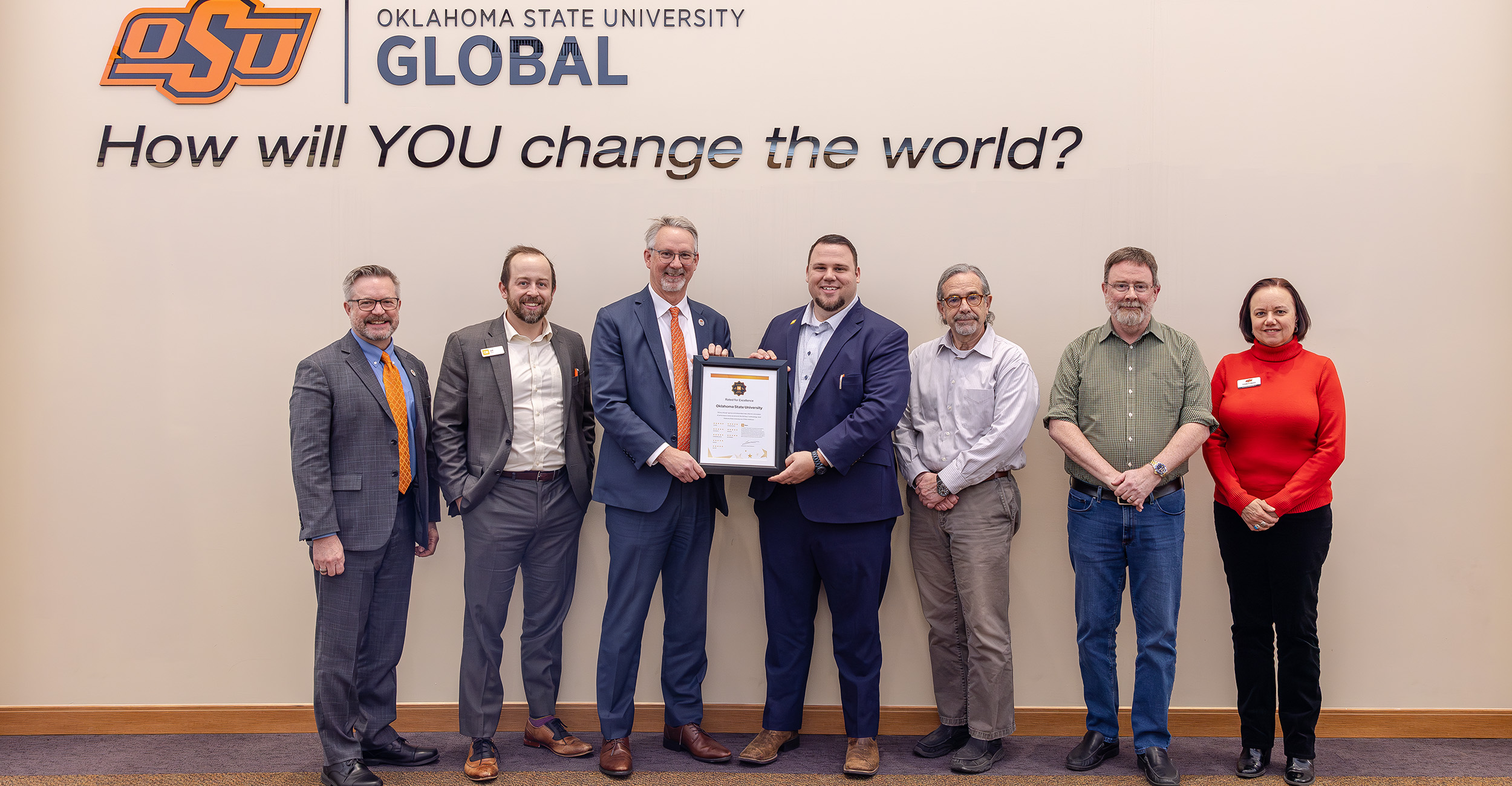 Seven people stand in front of a wall featuring the Oklahoma State University Global logo and the phrase 'How will YOU change the world?' One person in the center holds a framed certificate.