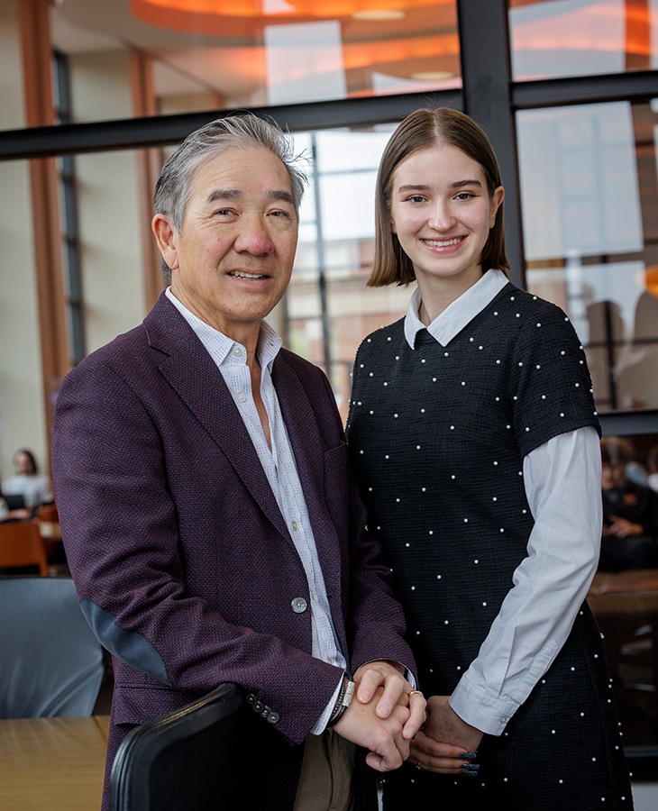 man and a woman stand side by side in an indoor setting with large windows and modern decor. The man wears a maroon blazer over a light blue shirt, and the woman wears a black dress with white polka dots over a white collared shirt. Both are smiling and looking at the camera.