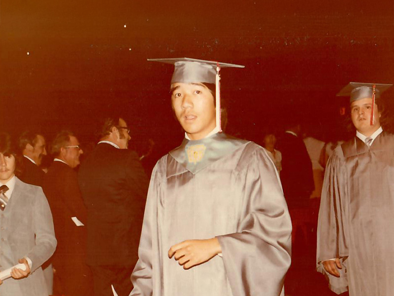 A person in a gray graduation gown and cap stands in the foreground holding a piece of paper. In the background, others in graduation attire and formal suits are visible in a dimly lit indoor venue, suggesting a graduation ceremony.