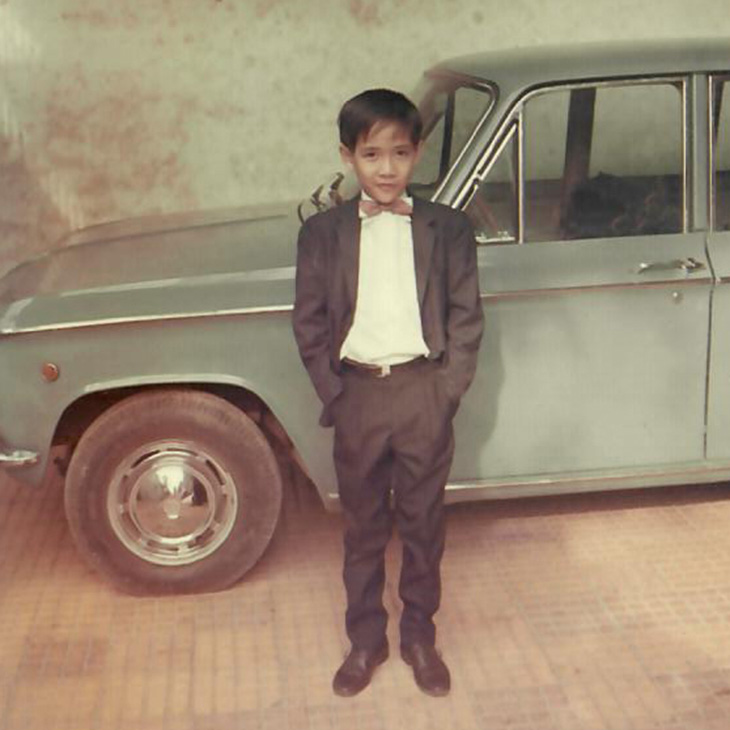 A young child wearing a formal suit and bow tie stands with hands in pockets on a tiled surface in front of a light blue vintage car with chrome details. The child has short hair and is looking directly at the camera. The car is parked against a plain wall.