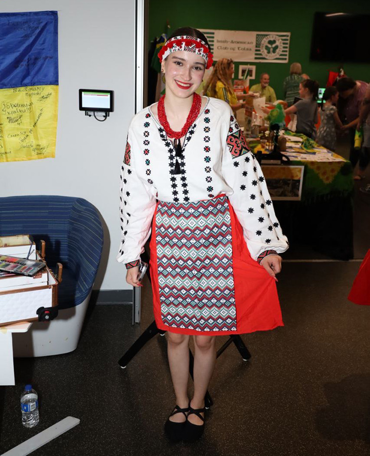 A person stands indoors wearing traditional Eastern European attire, including a white blouse with black and red embroidery, a red and blue patterned skirt, and a red headband with white decorations. They are holding a small object in one hand.