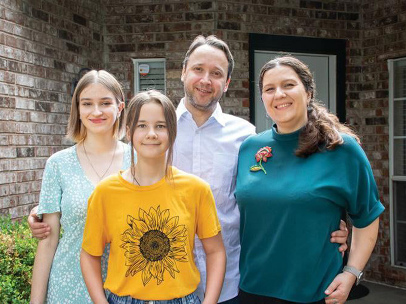 A family of four stands in front of a brick house. The group includes two adults and two children. One adult wears a light blue dress with white floral patterns, and the other wears a teal shirt with a red flower brooch. One child wears a yellow t-shirt with a large sunflower graphic, and the other wears a white shirt. They are smiling and have their arms around each other.