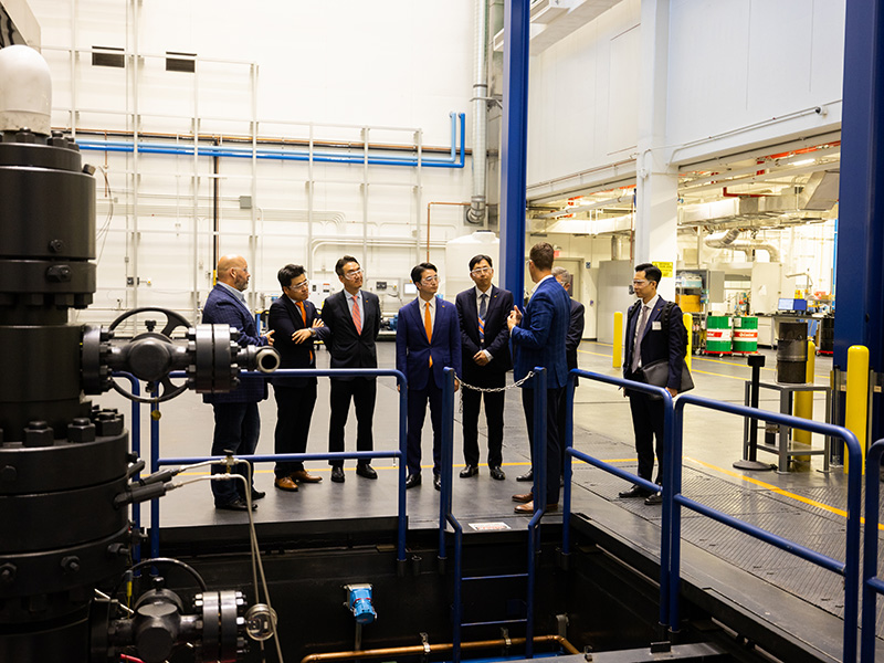 Seven individuals in business attire standing on a platform inside an industrial facility, engaged in discussion. One person is gesturing toward the group. The background includes industrial equipment, pipes, and machinery, suggesting a manufacturing or research setting.