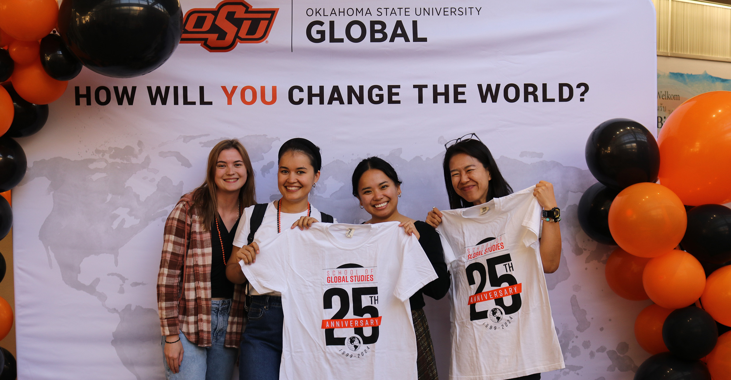Four individuals standing in front of a backdrop that reads 'OKLAHOMA STATE UNIVERSITY GLOBAL' and 'HOW WILL YOU CHANGE THE WORLD?' with a light gray world map. Two people in the center hold white T-shirts displaying 'GLOBAL STUDIES 25TH ANNIVERSARY' with a globe graphic.