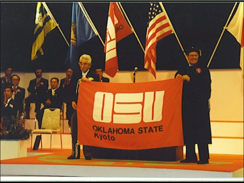 Two individuals on a stage holding a large red banner with white text that reads 'OSU OKLAHOMA STATE Kyoto.' Behind them are flags of the United States and Japan, with several seated attendees clapping in the background.