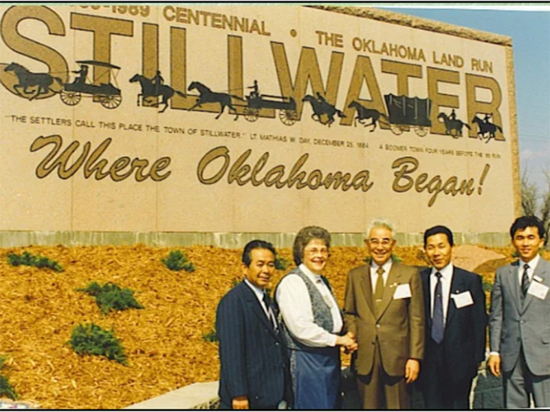 Five individuals in formal attire standing in front of a large commemorative sign that reads '1889–1989 Centennial The Oklahoma Land Run STILLWATER Where Oklahoma Began!' 