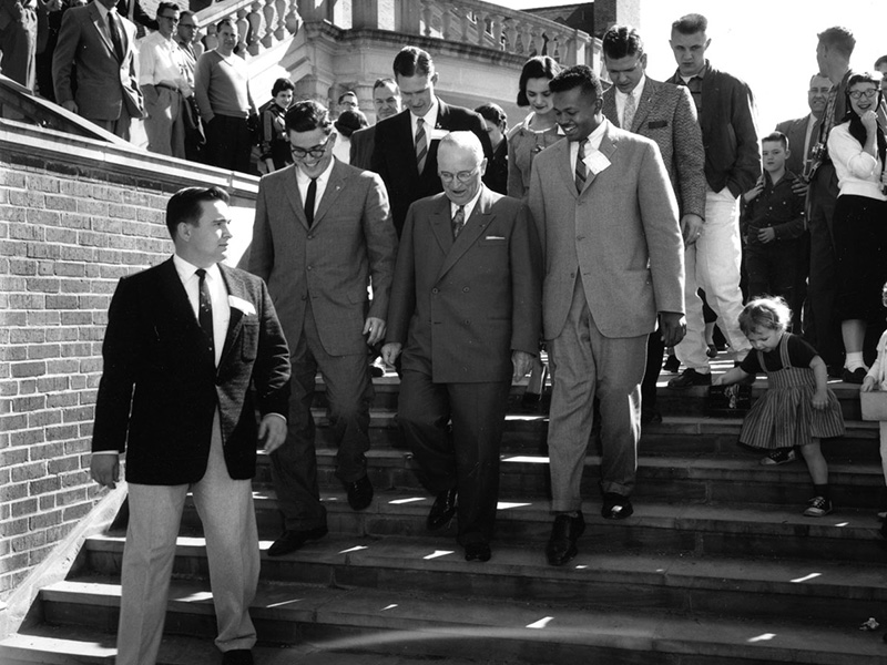 A group of people descending an outdoor staircase, including several men in suits and ties. An older man in a double-breasted suit and glasses is at the center, flanked by younger men in formal attire. Other individuals, some casually dressed, are also on the stairs. A small child in a dress stands at the bottom right corner. 