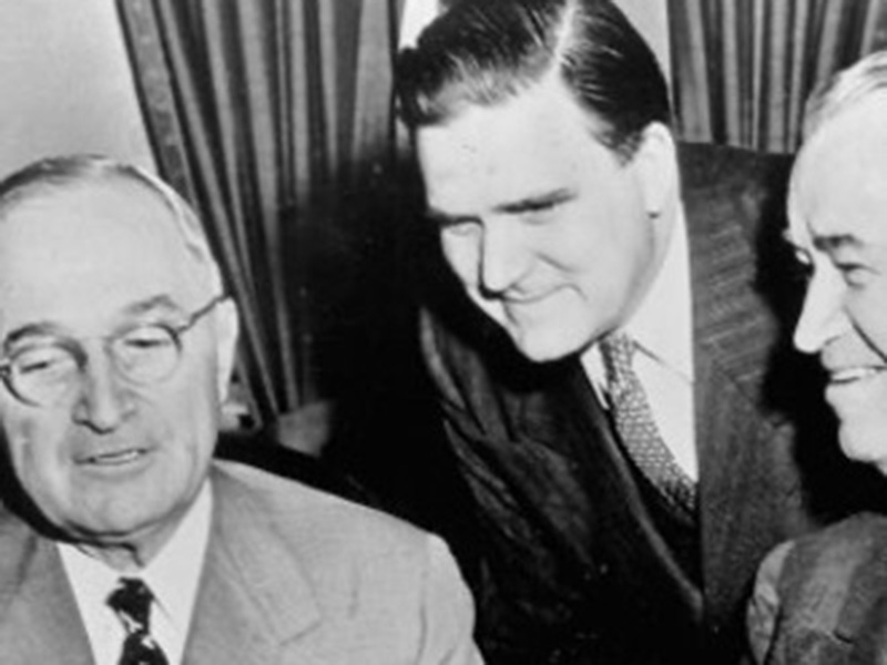 Three men in suits standing closely together in front of a curtain backdrop, engaged in conversation. The man on the left wears glasses and a light-colored suit with a patterned tie. The man in the middle, in a dark suit and tie, leans forward attentively. The man on the right, also in a dark suit, is smiling.