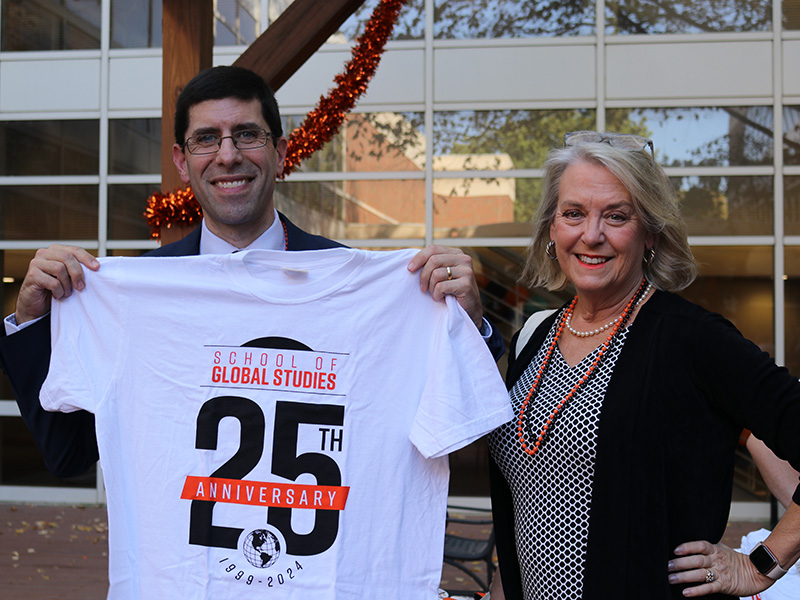 A man and a woman standing outdoors in front of a building with large windows. The man holds up a white T-shirt that reads 'School of Global Studies 25th Anniversary 1999–2024' in orange and black text. The woman, wearing a black cardigan and orange beaded necklaces, stands beside him smiling. An orange garland hangs in the background.