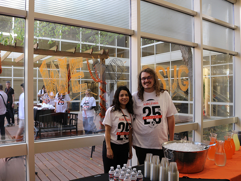 Two people standing indoors in front of a large window wearing matching white T-shirts with '25th Anniversary.'