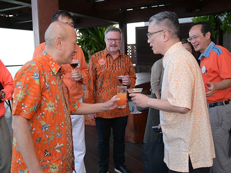 Six people stand together on a wooden deck under a covered outdoor area, surrounded by plants and wooden structures. They are dressed in orange-themed clothing, including patterned shirts and solid tops, and are holding drinks such as cocktails and juices.