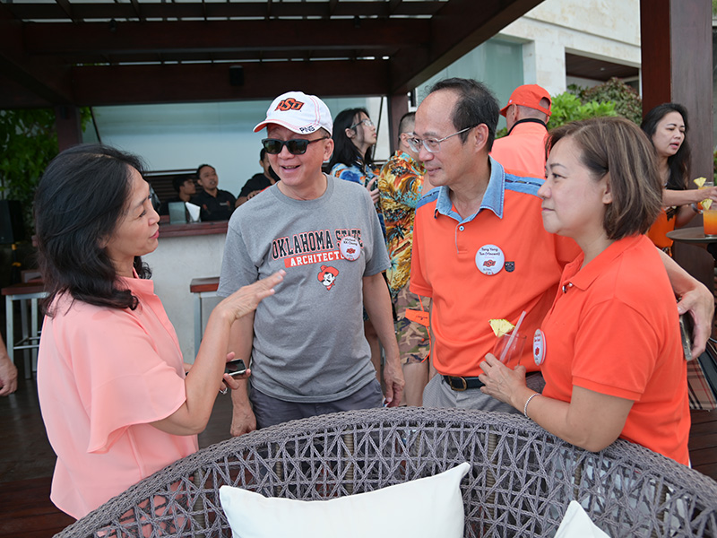 Four people are conversing at an outdoor event on a patio or deck area. One wears a gray Oklahoma State University t-shirt and a white cap, another wears an orange shirt with a name tag, and the others are dressed casually. 