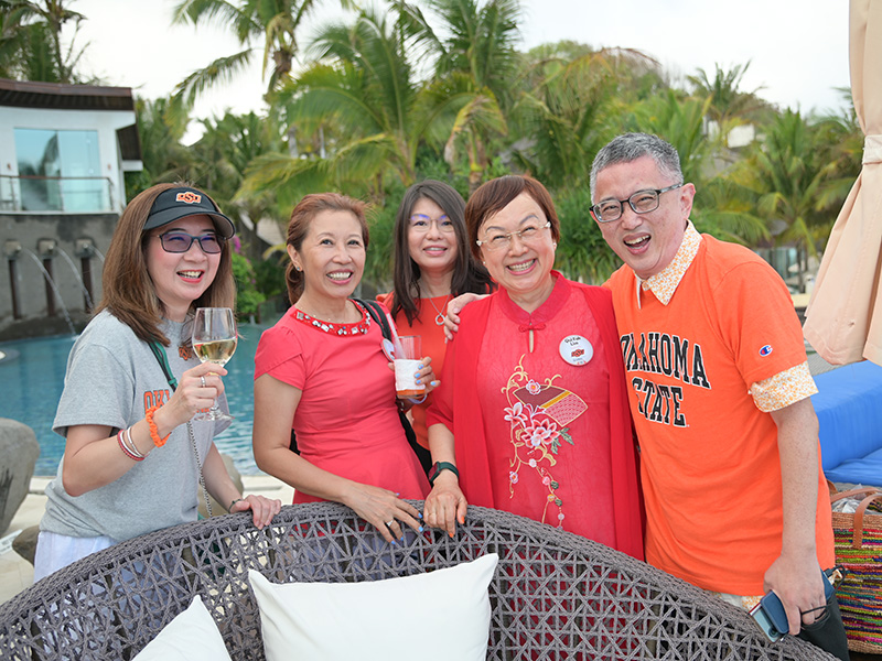 Five people stand together outdoors near a pool with palm trees in the background. They are smiling and appear to be enjoying themselves. One person holds a glass of white wine, another has a drink with ice, and one wears an orange 'Oklahoma State' shirt.