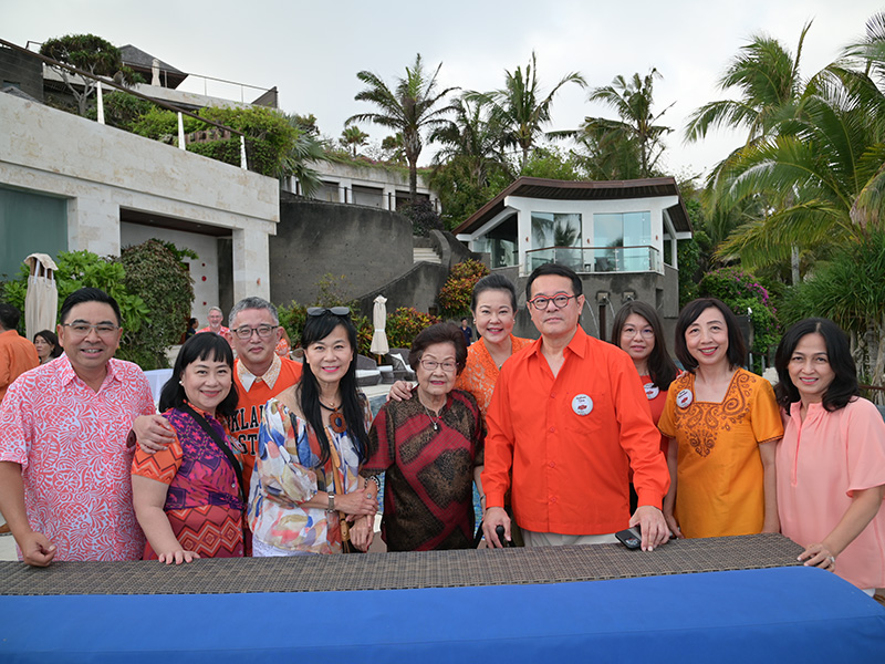 Ten people stand together outdoors in front of a modern house, surrounded by palm trees and greenery. They are dressed in colorful clothing, mainly in orange and pink tones, and are smiling at the camera. 