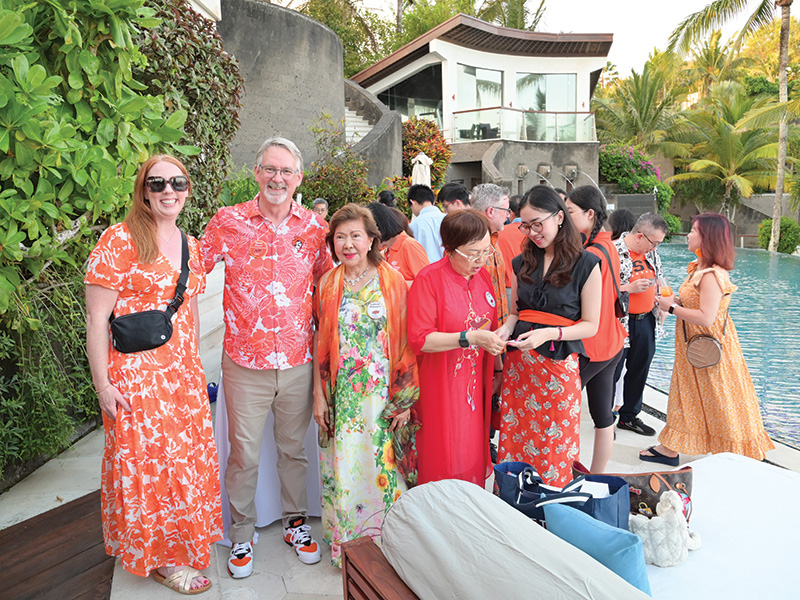  group of people gathered outdoors near a pool and modern house, surrounded by lush greenery. They are dressed in colorful clothing, mainly in orange and red with floral patterns. Some individuals are smiling at the camera, while others are conversing or looking at items in their hands.