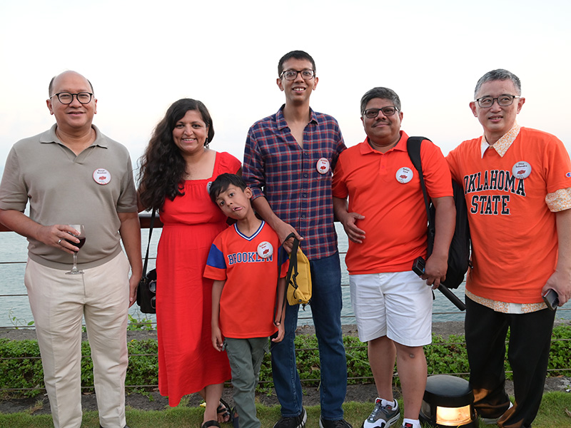 x people stand outdoors near a body of water with greenery and a railing in the background. The group includes four adults and two children. One adult wears a beige polo shirt and holds a glass, another is in a red dress, one wears a blue checkered shirt, and another wears an orange 'Oklahoma State' t-shirt.