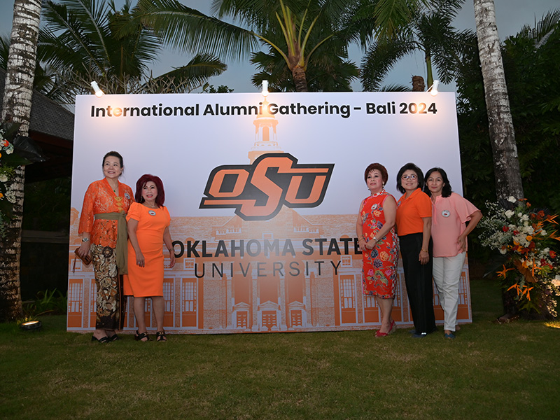 Five people stand in front of a large sign that reads 'International Alumni Gathering – Bali 2024' featuring the Oklahoma State University logo and a building image. The event is held outdoors, with palm trees and greenery in the background.