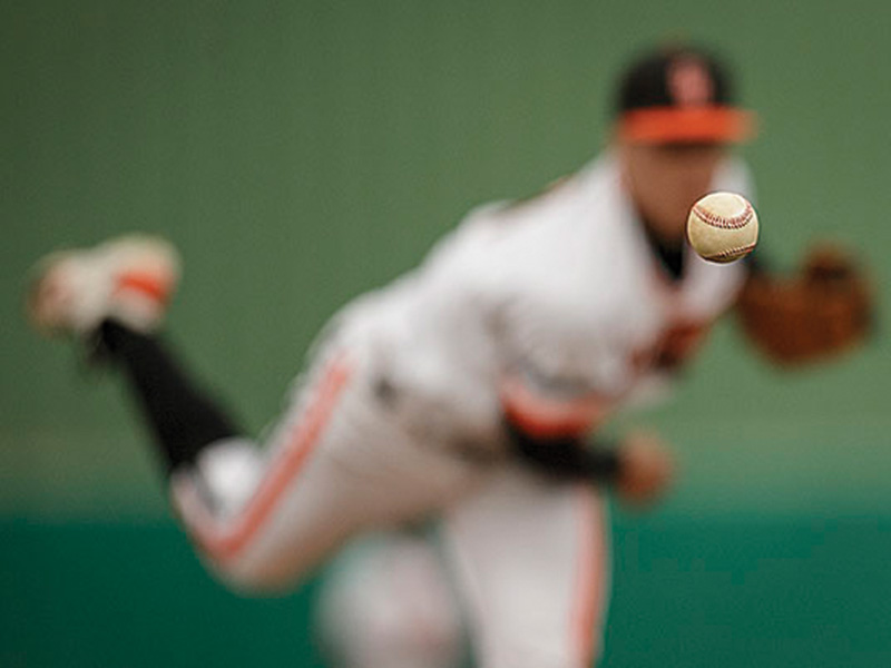 OSU baseball A baseball pitcher in a white uniform with orange and black accents is captured mid-pitch. The baseball is in sharp focus as it moves toward the camera, while the pitcher’s body is blurred in the background to emphasize speed and motion.