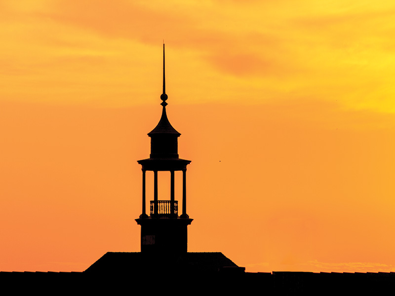 Stillwater Sunrise A silhouette of a building with a tall, pointed spire set against a vibrant orange and yellow sunset sky. The building features an open structure near the top with columns and a small railing.