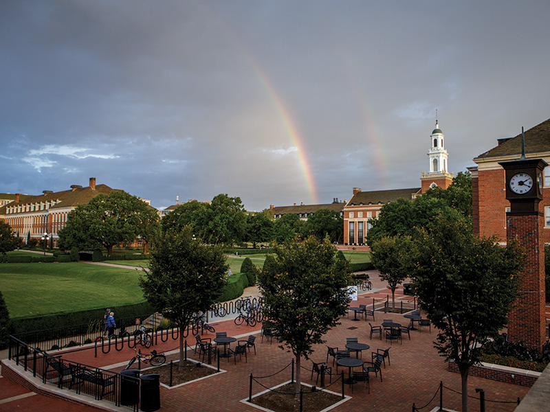 Double Rainbow A scenic view of a university campus with a brick courtyard featuring tables and chairs, surrounded by trees. Large red brick academic buildings with white trim stand in the background, and a clock tower is visible on the right. A double rainbow arcs across the cloudy sky above.