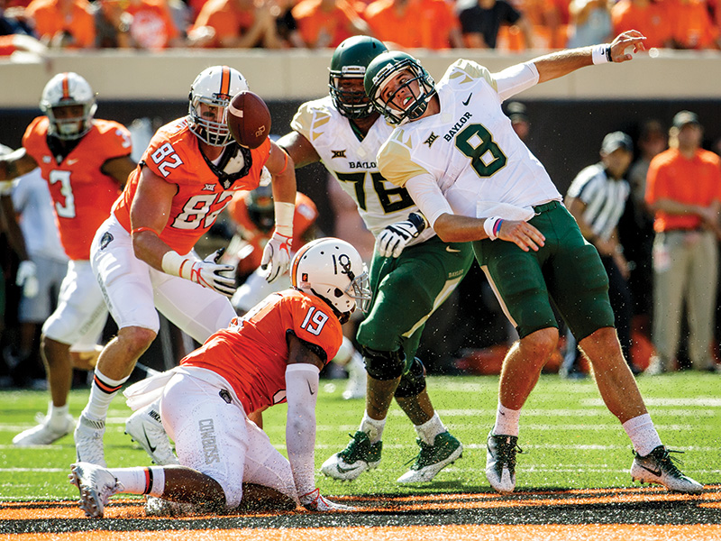 OSU football A college football game between Baylor and Oklahoma State. Oklahoma State player number 19 is on the ground while teammate number 82 reaches for Baylor player number 8. Baylor player number 76 is also visible.