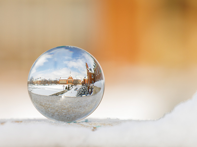 Ornament A clear glass sphere resting on snow reflects an upside-down image of a snow-covered campus with red brick buildings and a central clock tower. The background is blurred, highlighting the sharp reflection inside the sphere.