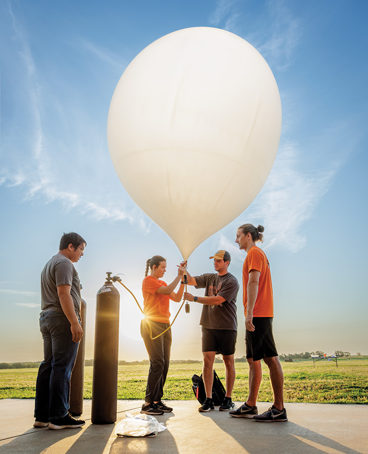 Balloon Launch Four individuals stand on a concrete surface outdoors, preparing to launch a large white weather balloon tethered to a gas cylinder. The balloon is partially inflated. The sky is clear with wispy clouds, and the low sun casts long shadows.