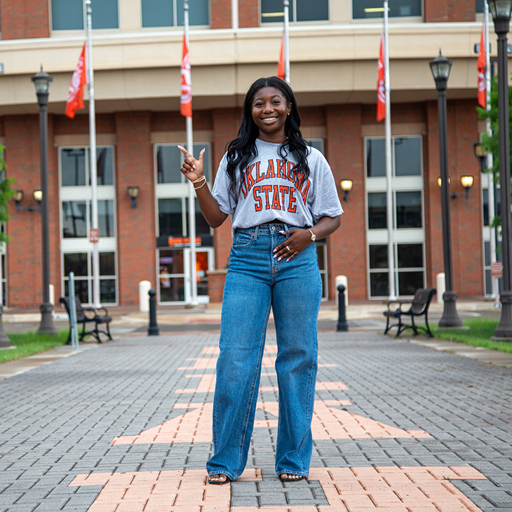 A woman wearing a gray "Oklahoma State" T-shirt and blue jeans stands on a paved walkway in front of a building with large windows and multiple orange flags.