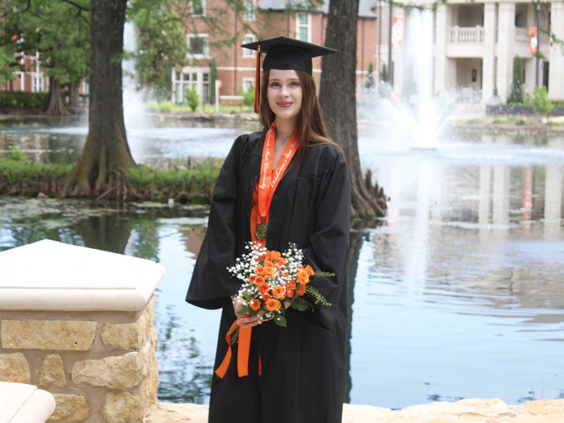 A woman in a black graduation gown and cap stands in front of a pond with fountains, holding a bouquet of orange and white flowers tied with an orange ribbon. 