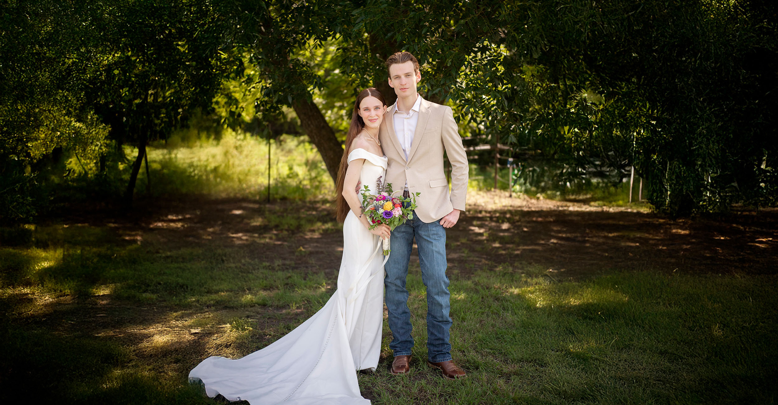 A couple stands outdoors under tree shade. A woman wears a white wedding dress and holds a colorful bouquet. A man wears a beige blazer, white shirt, blue jeans, and brown shoes. They stand on grass with sunlight filtering through the trees.