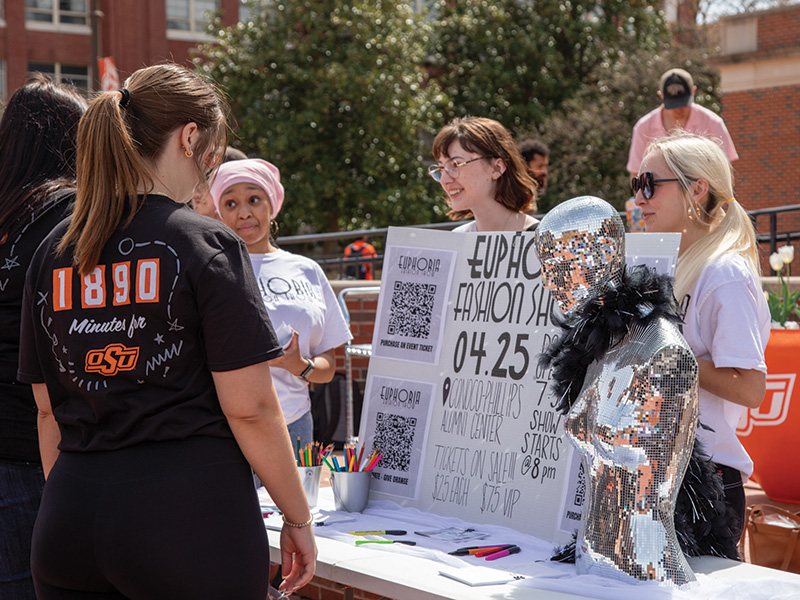 A group of people gathered around a table at an outdoor event featuring colorful markers and papers. A mannequin covered in reflective material and wearing a black feather boa stands beside the table. 