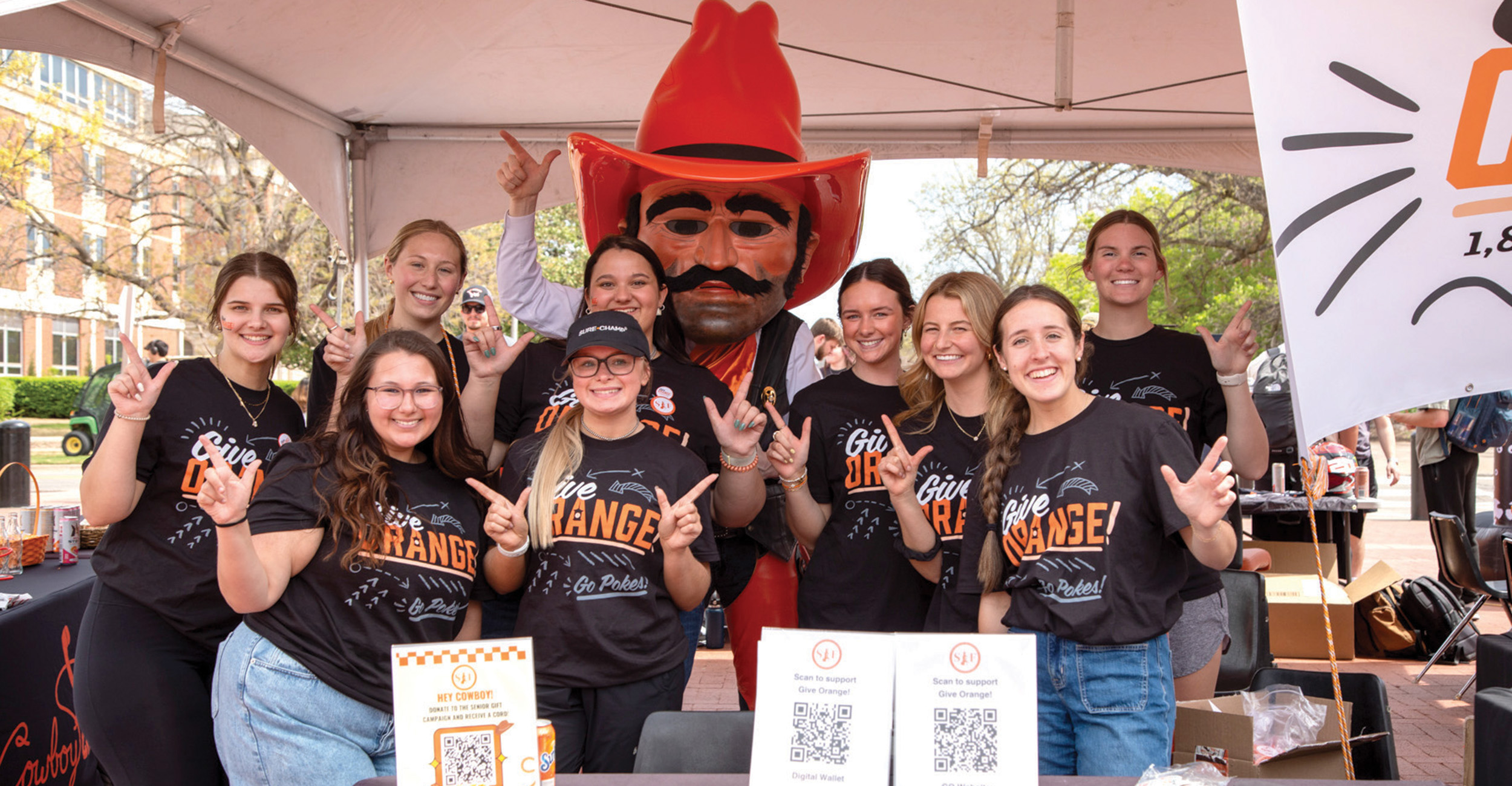 A group of people wearing matching black 'Give Orange' T-shirts pose under a tent at an outdoor event. They stand around a table with QR code signs, making hand gestures.