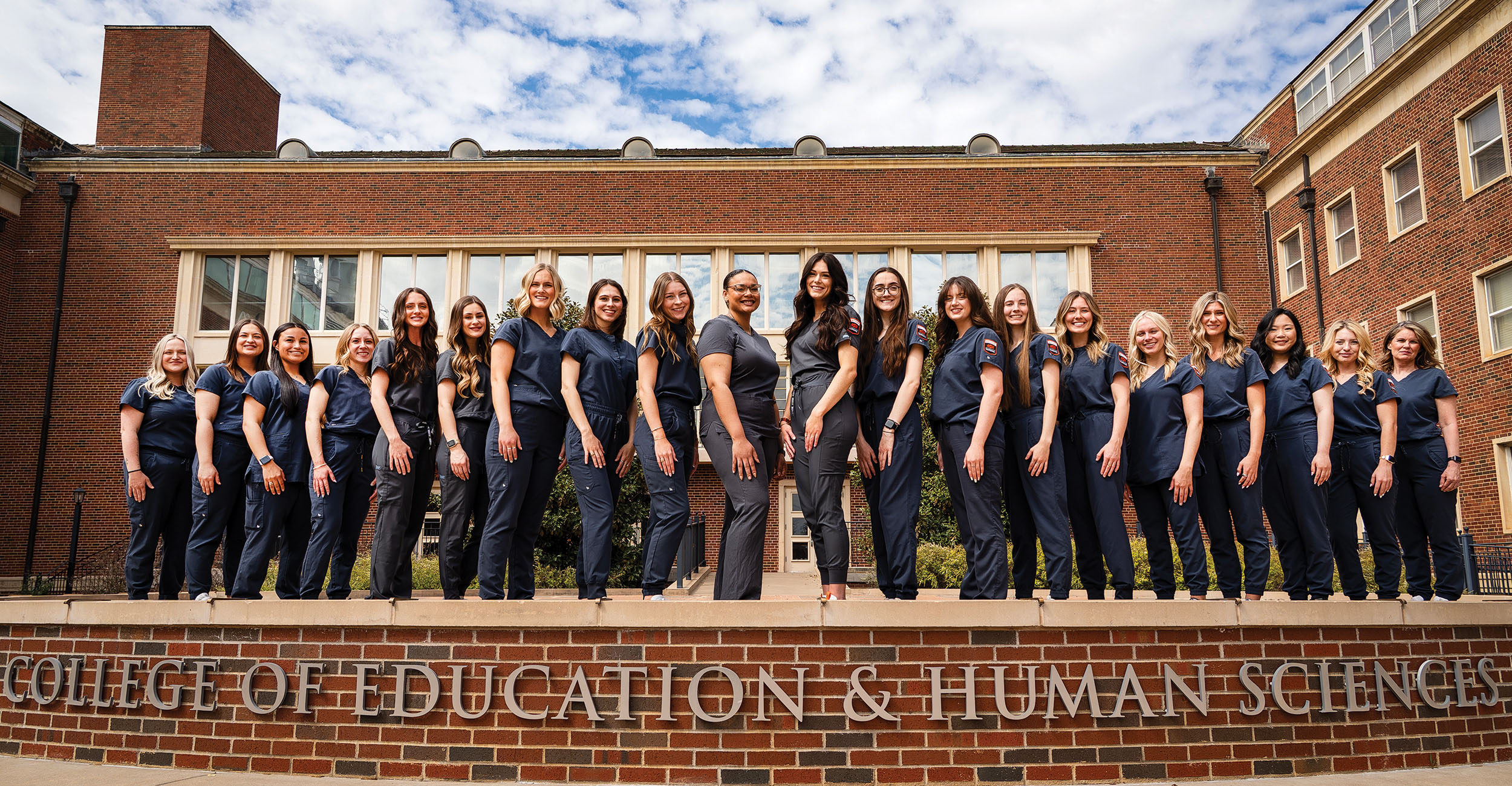 A group of people in matching dark blue uniforms stand in a line on a brick platform in front of a building labeled 'College of Education & Human Sciences.'