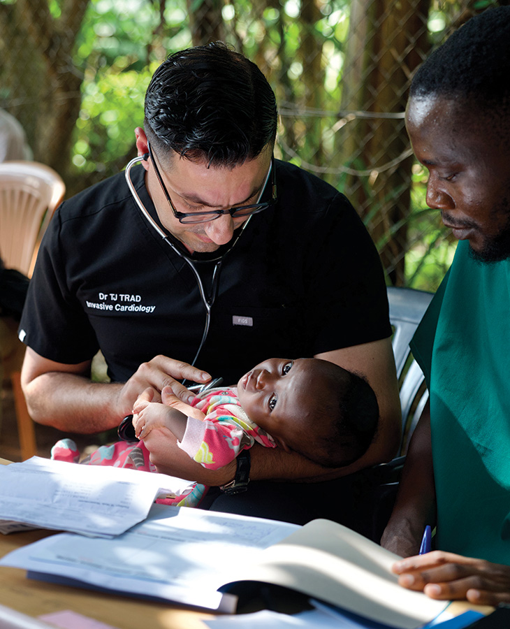 A doctor with a name tag reading 'Dr. TJ Trad, Invasive Cardiology' holds a baby dressed in a colorful outfit while seated at a table with open books and papers, suggesting a medical consultation.