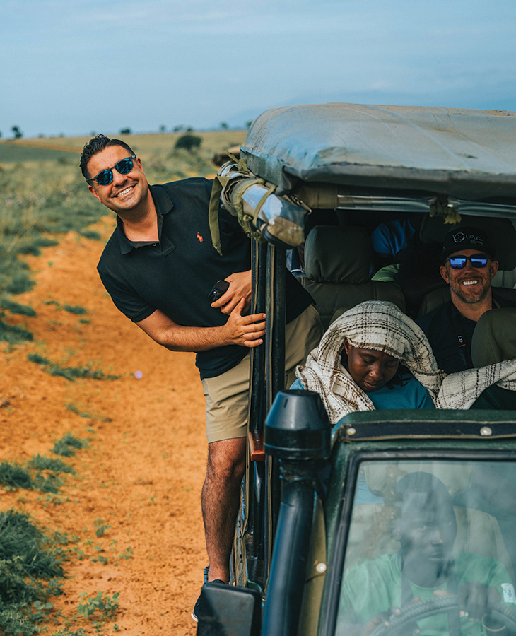 A group of people on a safari ride in an open-top vehicle, with one person standing and leaning out of the side while holding onto the frame. The surrounding landscape features dry terrain, patches of grass, and a dirt road extending into the distance.
