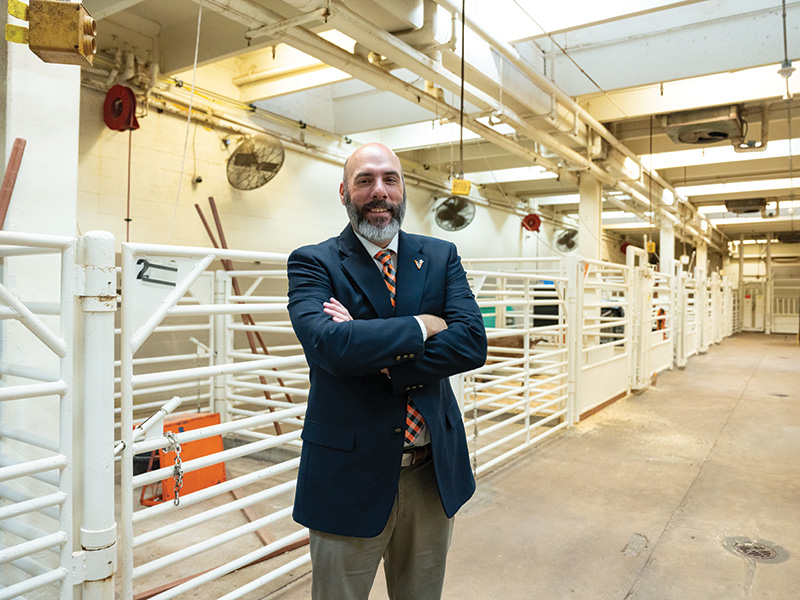 A person in a suit stands with arms crossed in a brightly lit industrial hallway featuring white metal railings, pipes, fans, and various machinery in the background.