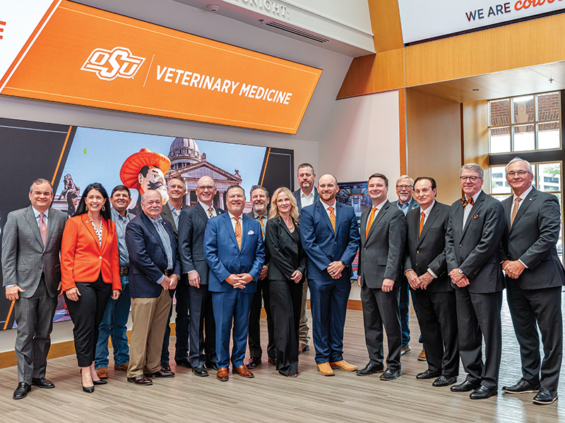 A group of people in formal attire stand in a lobby area with large digital screens behind them. One screen displays 'OSU VETERINARY MEDICINE' and an image of a statue wearing an orange hat. Another screen shows part of a building. 