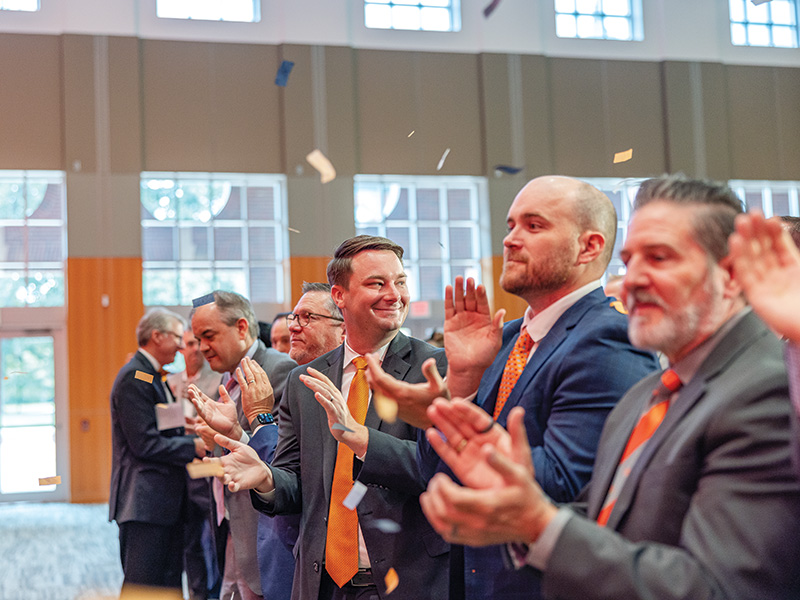 A group of people in business attire, including suits and ties, stand in a large room with tall windows, clapping as confetti falls from above. The background features a wooden panel wall and natural light streaming through the windows.