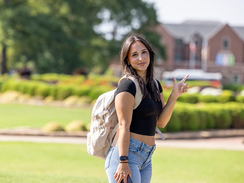 A woman with long hair stands outdoors on a sunny day, wearing a black crop top, light blue jeans, and a beige backpack.
