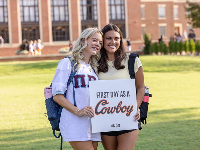 Two people stand on a grassy area in front of a brick building with large windows, holding a sign that reads 'FIRST DAY AS A Cowboy.'