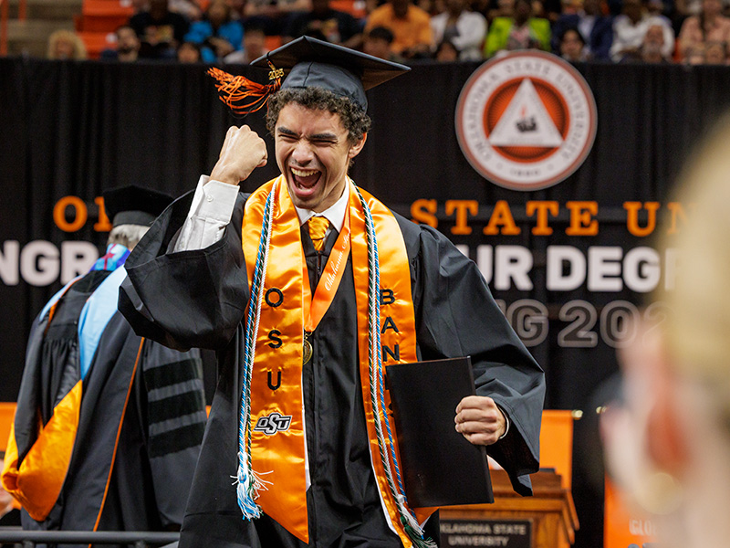 graduate in a cap and gown stands on stage, raising a fist in celebration while holding a diploma. The graduate wears an orange stole with 'OSU' and 'BAND' written on it, along with several cords.