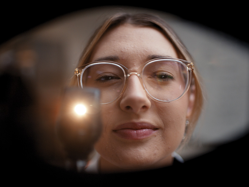 A person’s face is centered in the image, surrounded by a dark vignette-like border. The setting appears to be indoors with soft, indistinct lighting in the background."