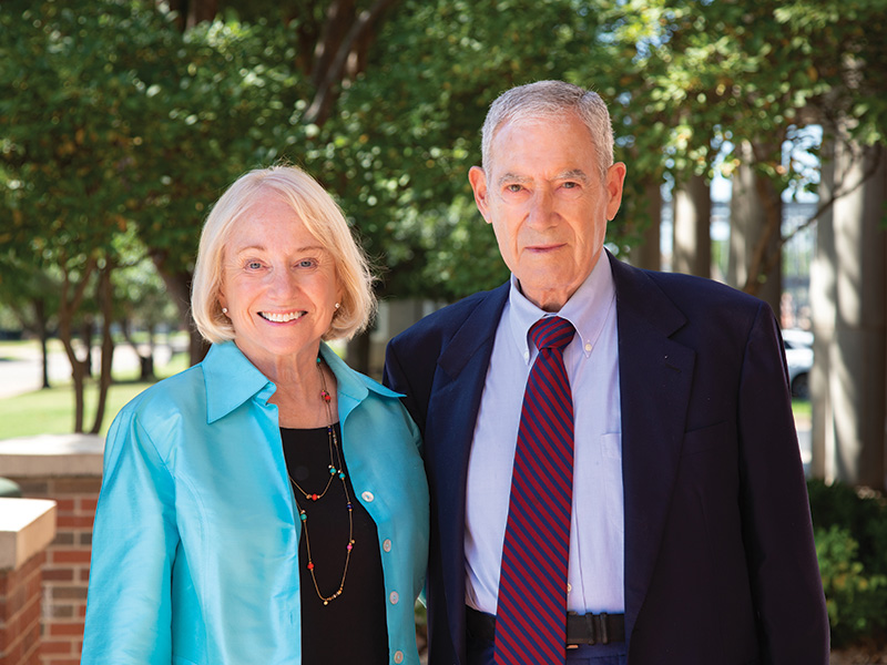 An elderly couple stands outdoors in front of trees and a brick structure. The woman wears a turquoise jacket over a black top with a necklace, and the man is dressed in a dark suit with a light blue shirt and red striped tie.