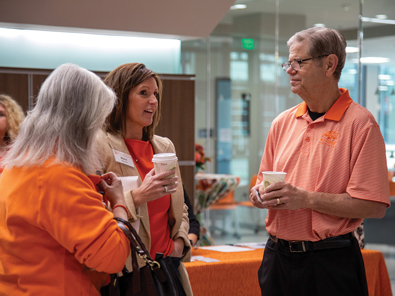 Three people stand and converse indoors, each holding a cup of coffee. 