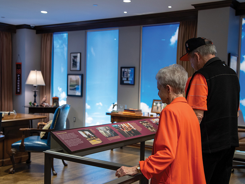 Two elderly individuals view an exhibit in a museum or gallery. The exhibit features a display case with informational panels and photographs.
