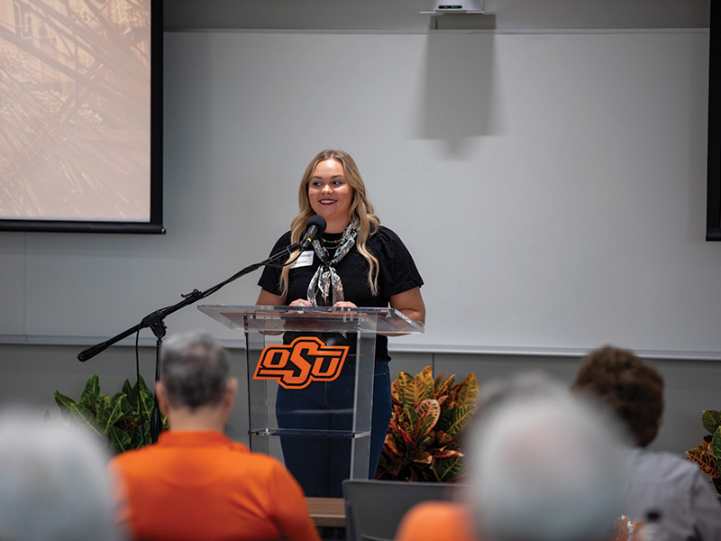 A person stands behind a clear podium with the Oklahoma State University (OSU) logo, speaking into a microphone. The speaker wears a black shirt and a scarf. 