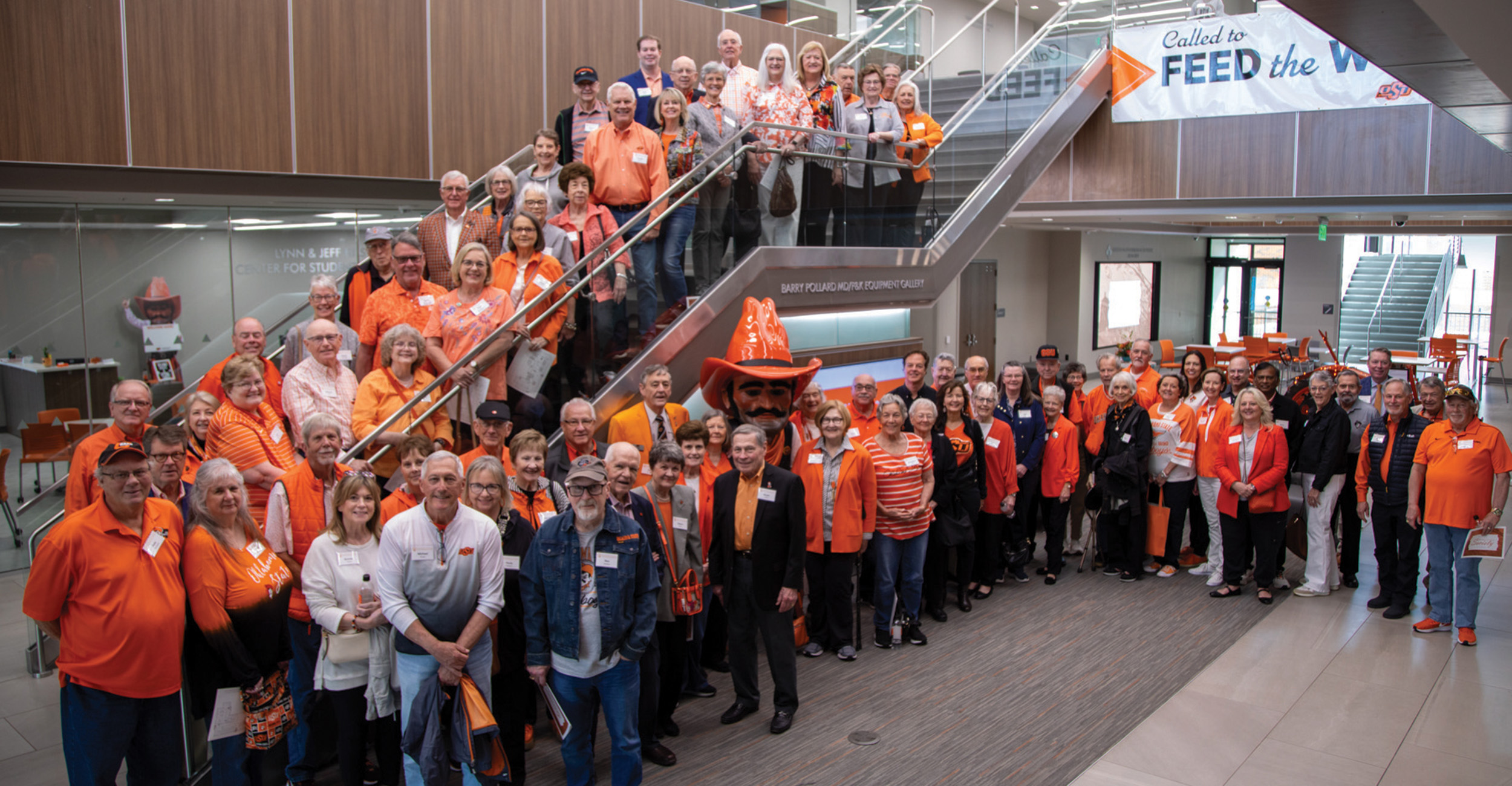 A large group of people, many dressed in orange, gather indoors around a staircase in a modern building.